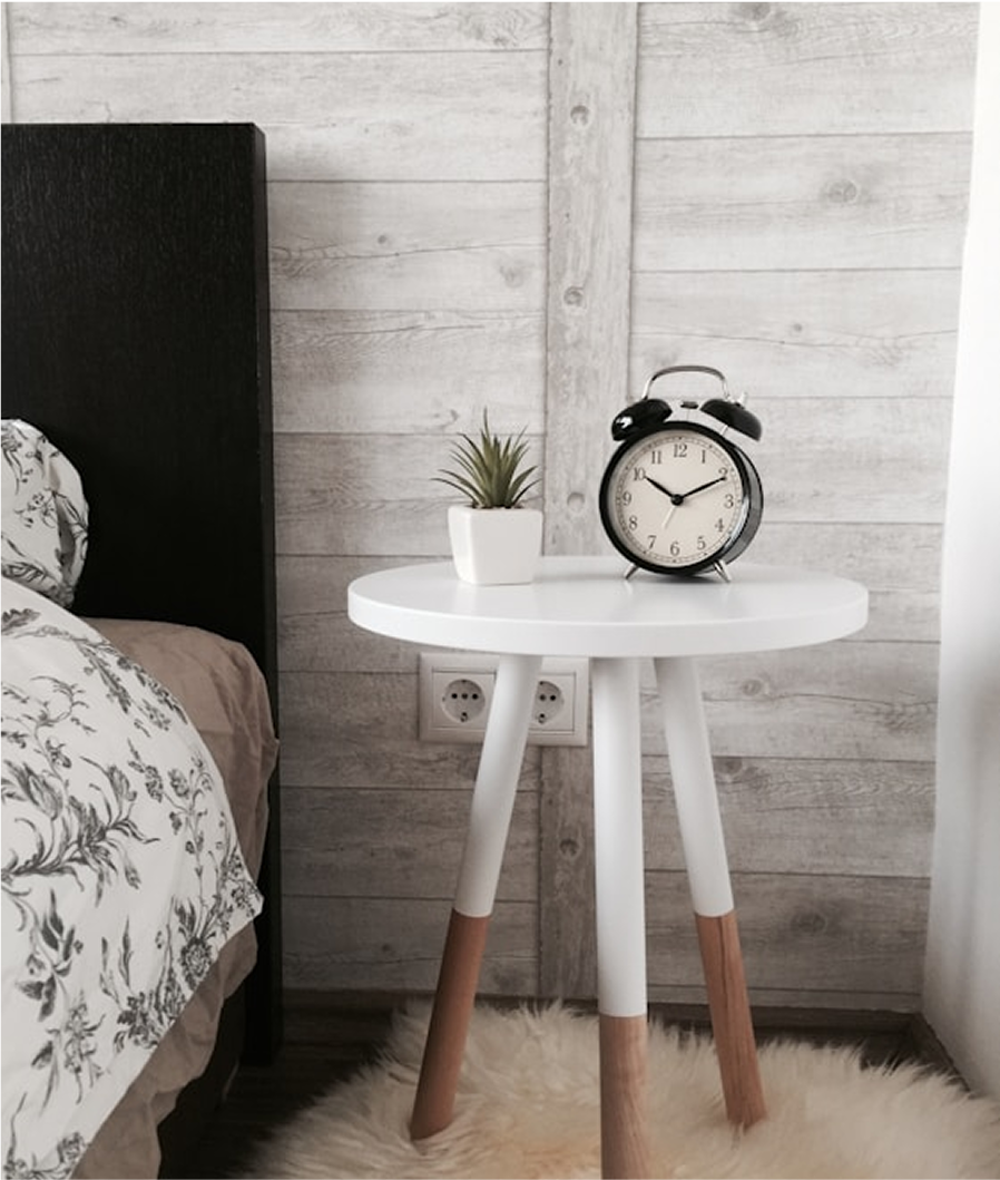 A modern white bedside table with wooden legs, featuring a classic black alarm clock and a small potted plant.